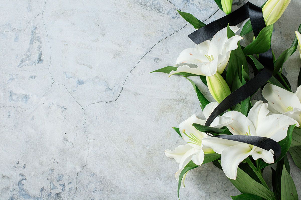 White lilies with a black ribbon on a gray textured background, likely for a memorial.