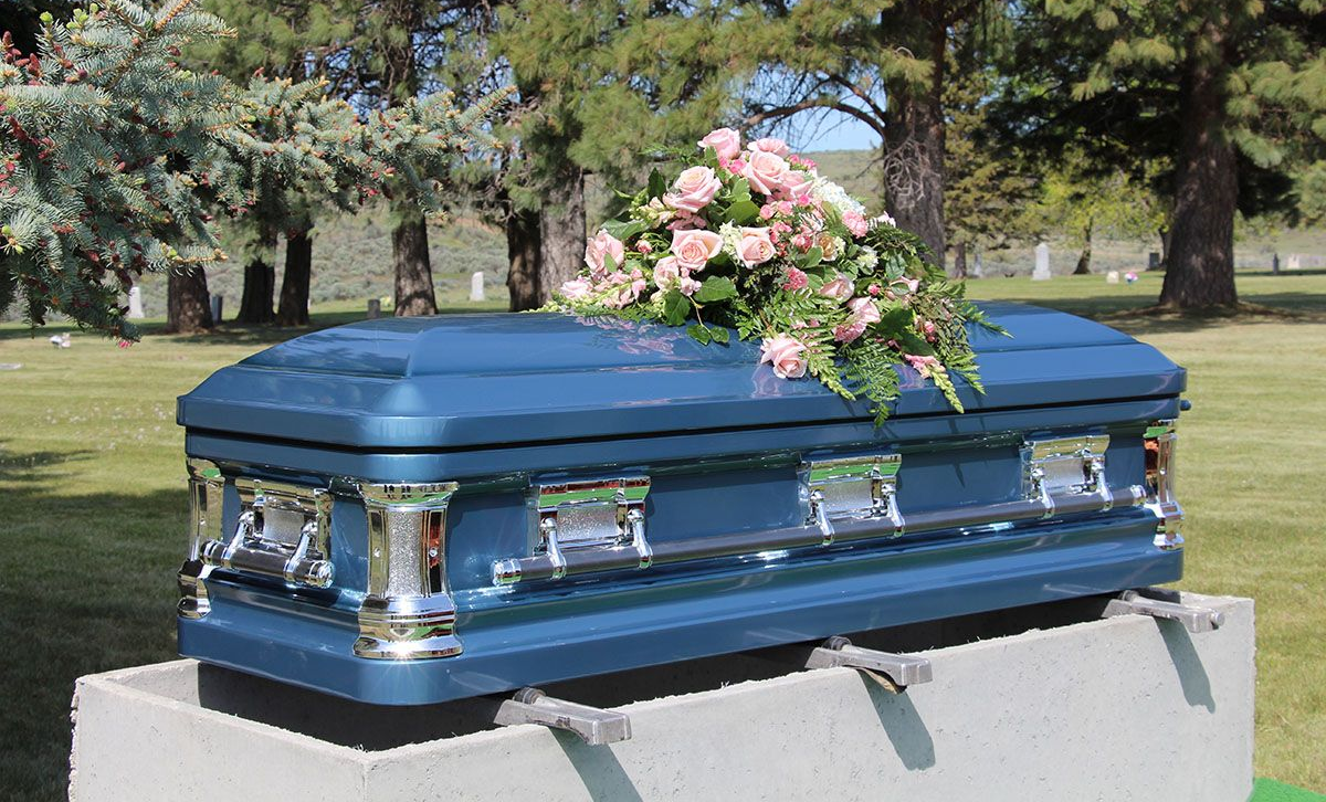 Blue casket with floral arrangement, outdoors on a stone platform in a cemetery.
