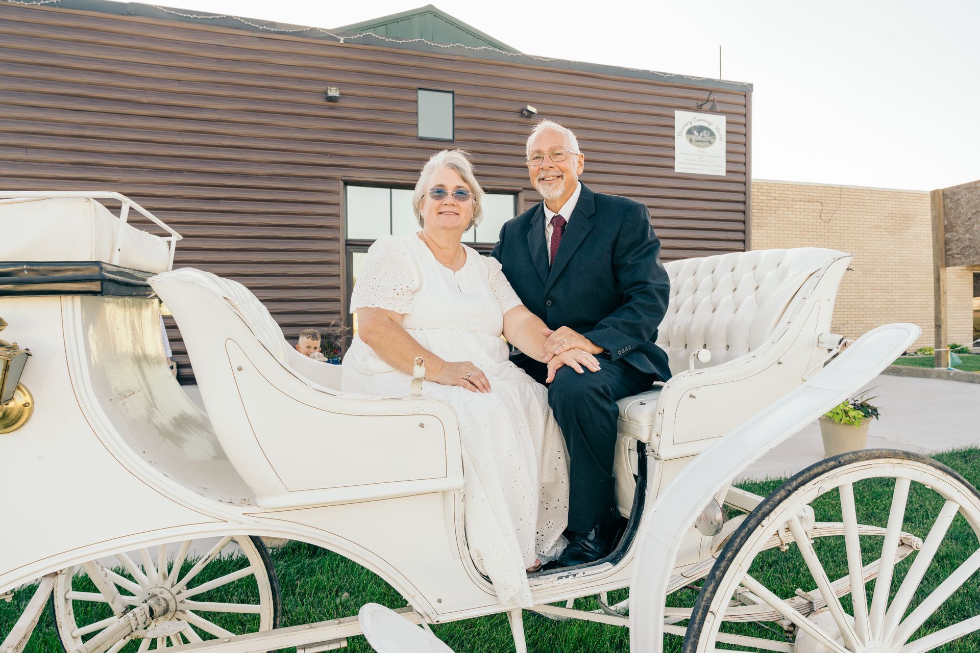 A couple sits in a white horse-drawn carriage parked in front of a dark horizontal-sided building. A couple sits in a white horse-drawn carriage parked in front of a dark horizontal-sided building.
