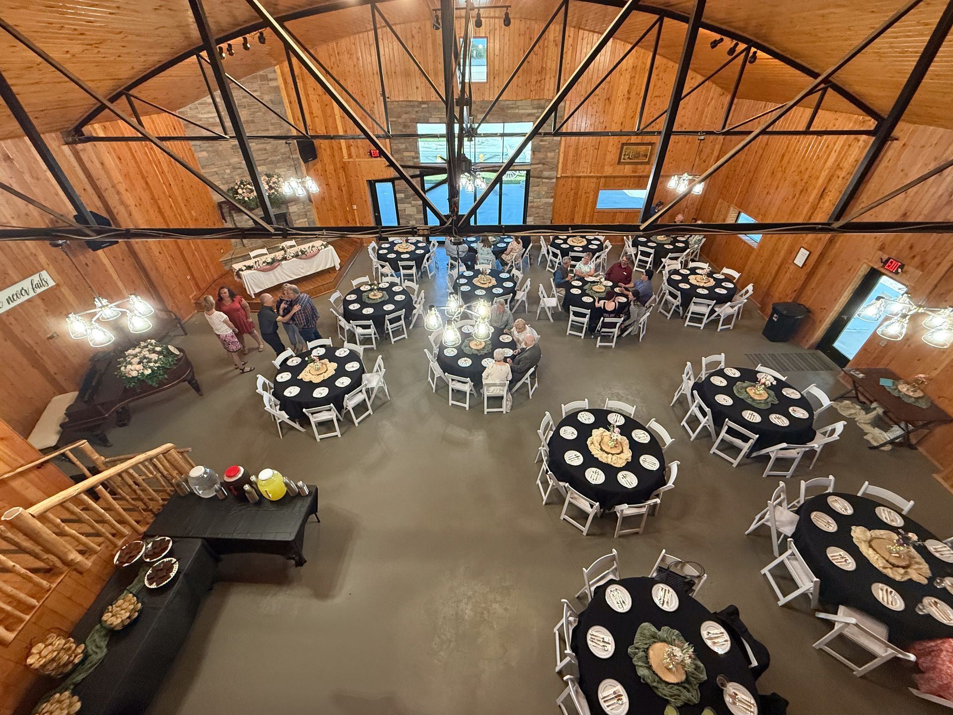 High-angle view of a rustic event hall with wooden walls, black-clothed round tables, and people gathering.