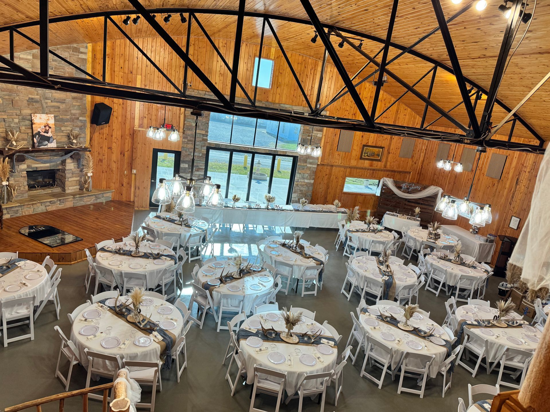 High-angle view of a rustic event hall set up with round tables for a dinner, featuring wooden walls and a stone fireplace.
