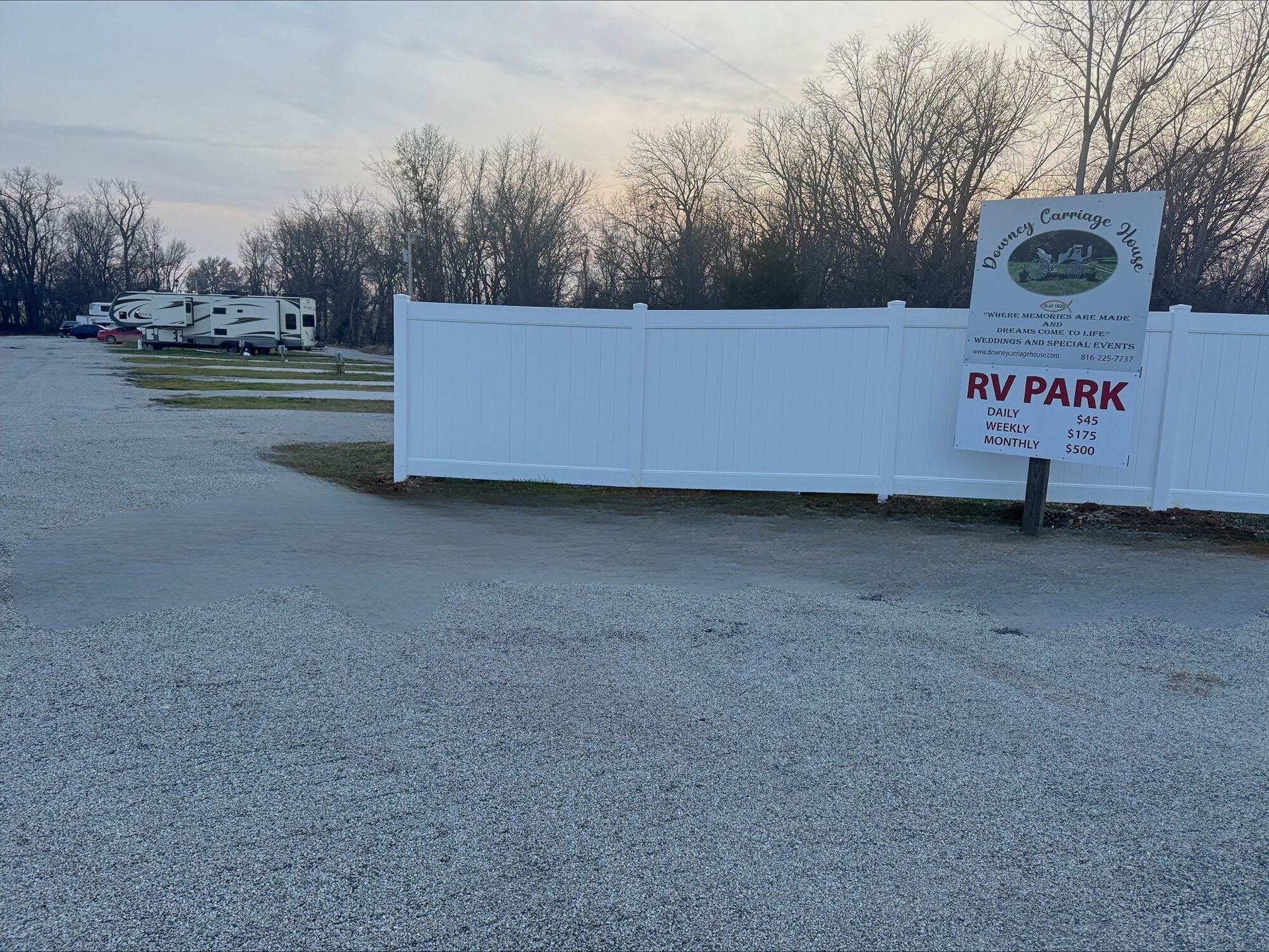 Gravel RV park with white fence and sign; RVs parked in the distance, trees and overcast sky. Gravel RV park with white fence and sign; RVs parked in the distance, trees and overcast sky.
