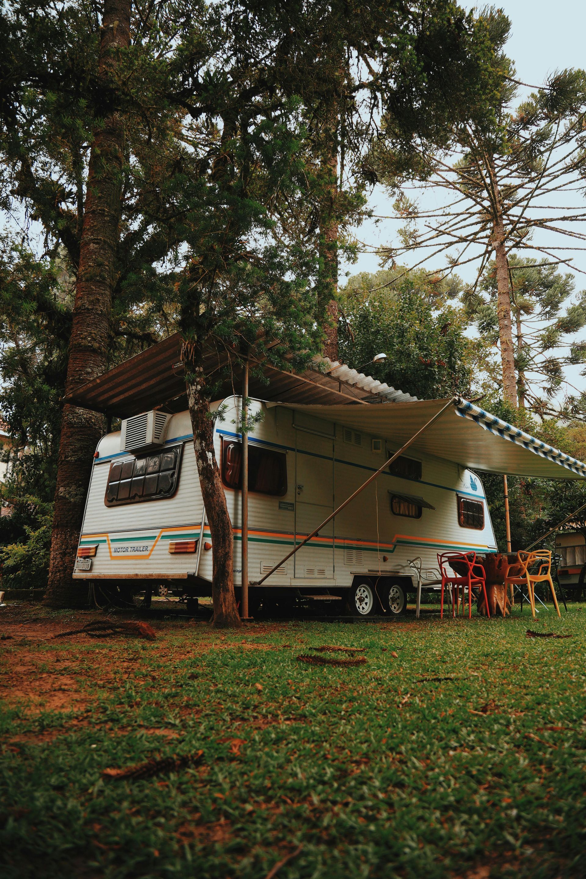 A white camper trailer with awning set up in a grassy area among tall trees. A white camper trailer with awning set up in a grassy area among tall trees.