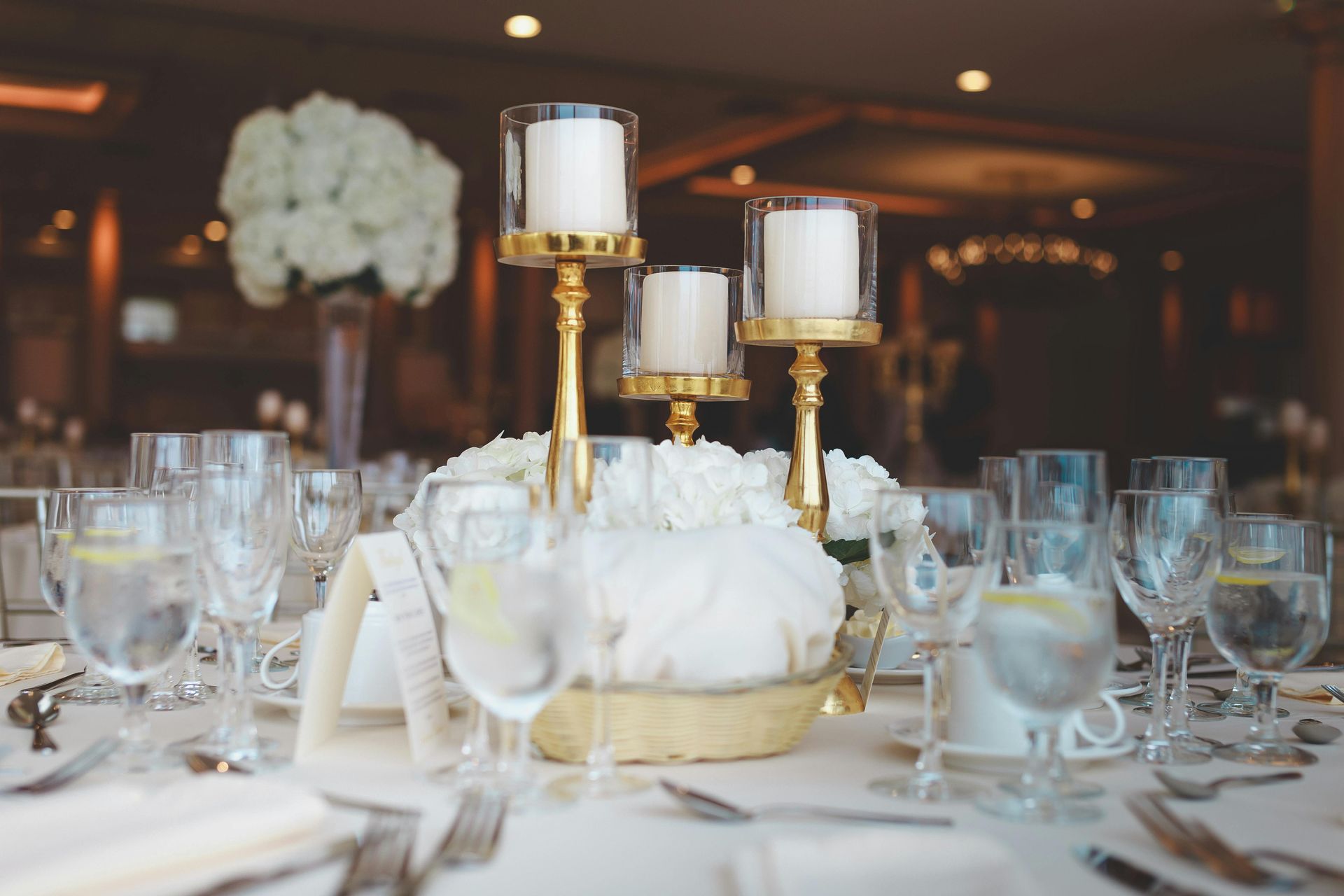 Elegant wedding table setting with gold candelabras, white flowers, and glassware in a well-lit dining room.