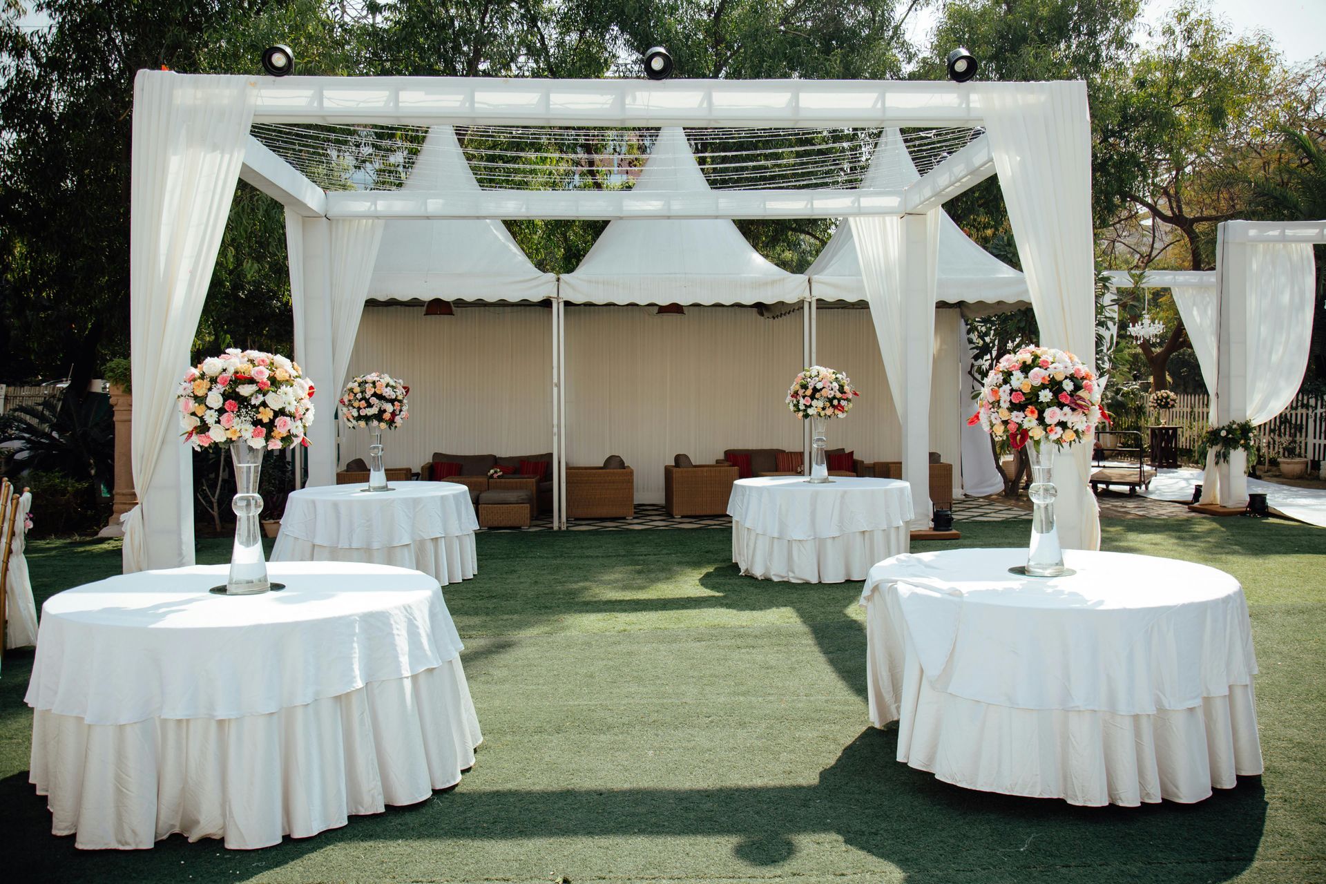 Outdoor event setup with round tables, floral arrangements, and white tent structures on grass.