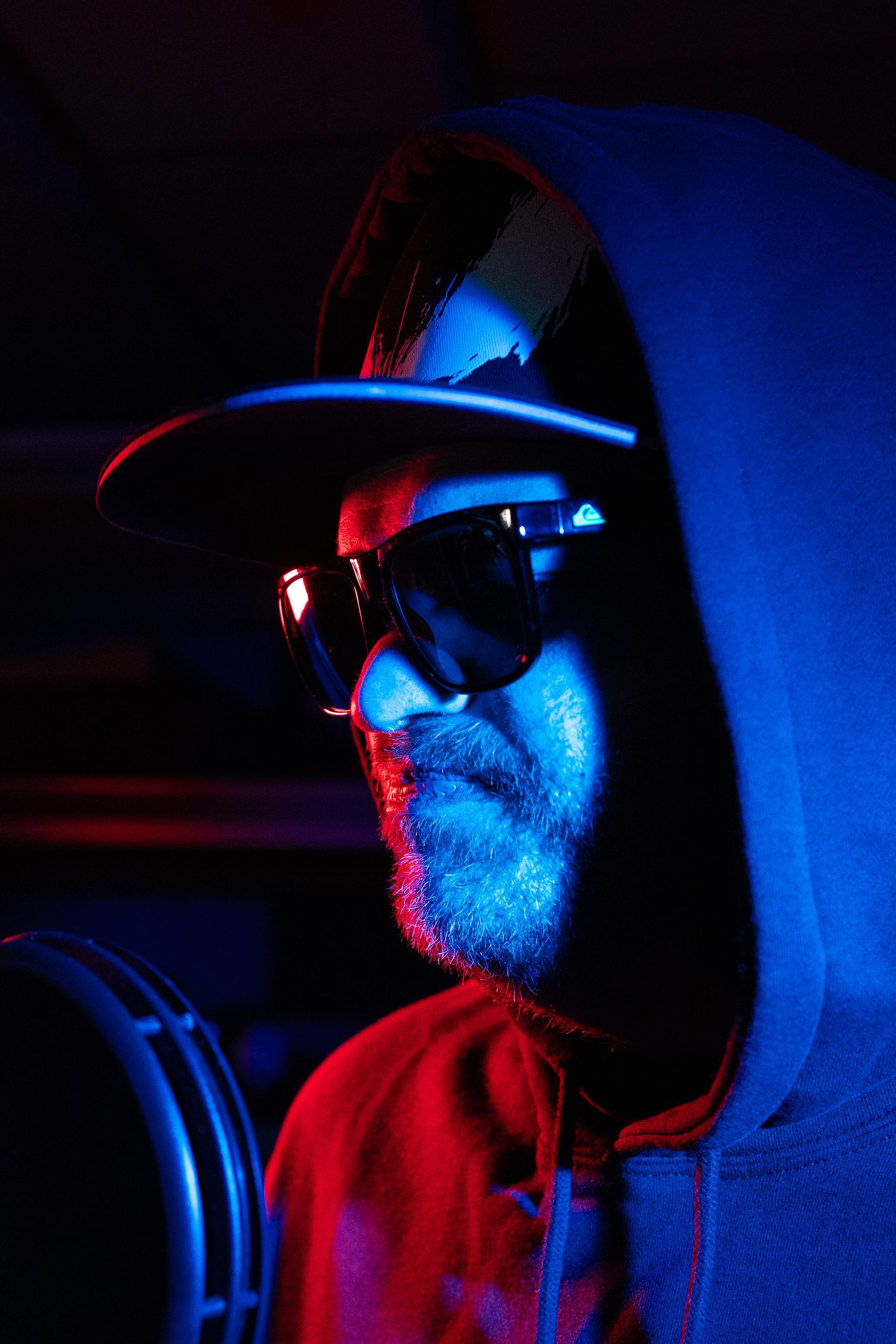 A man wearing sunglasses and a hat is standing in front of a microphone in a dark room.