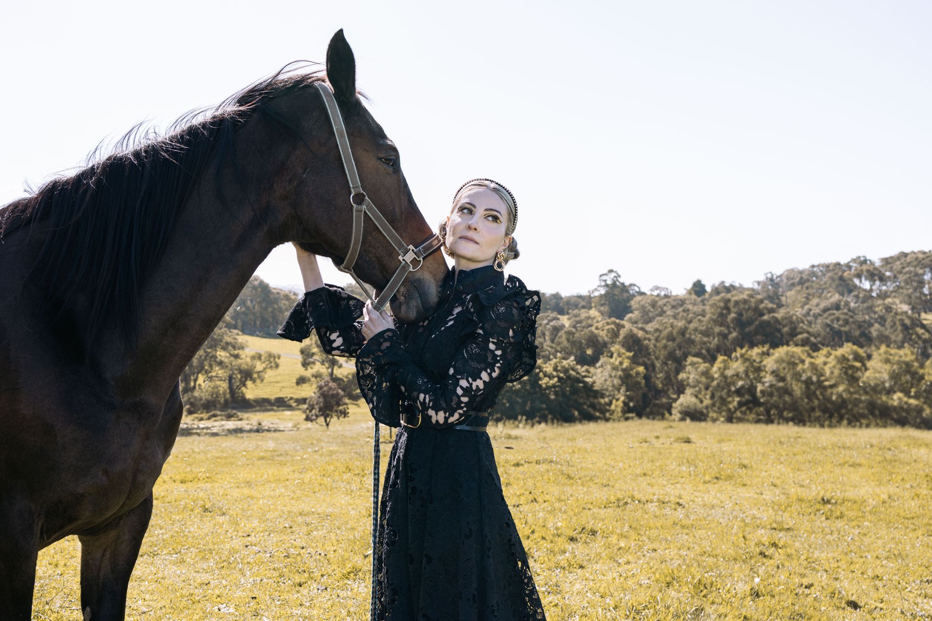 A woman in a black dress is standing next to a black horse in a field.