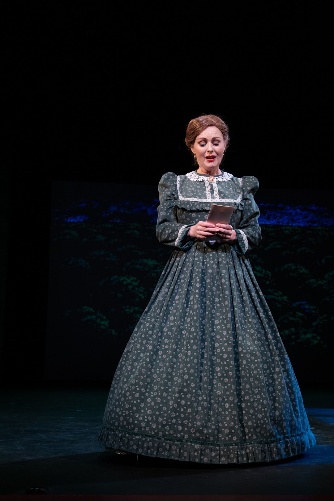 A woman in a long dress is standing on a stage holding a book.