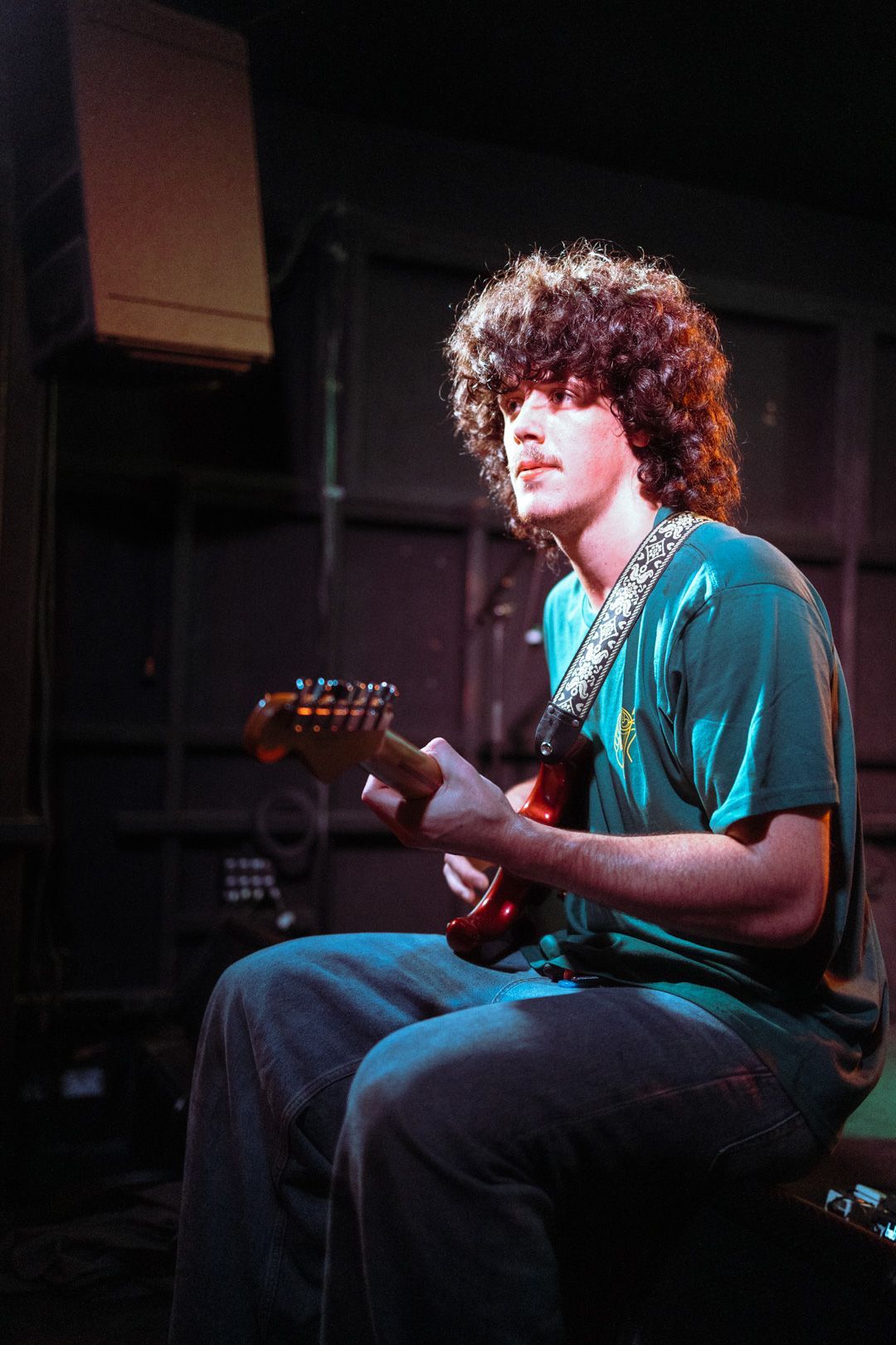 A young man with curly hair is sitting on a stage playing a guitar.