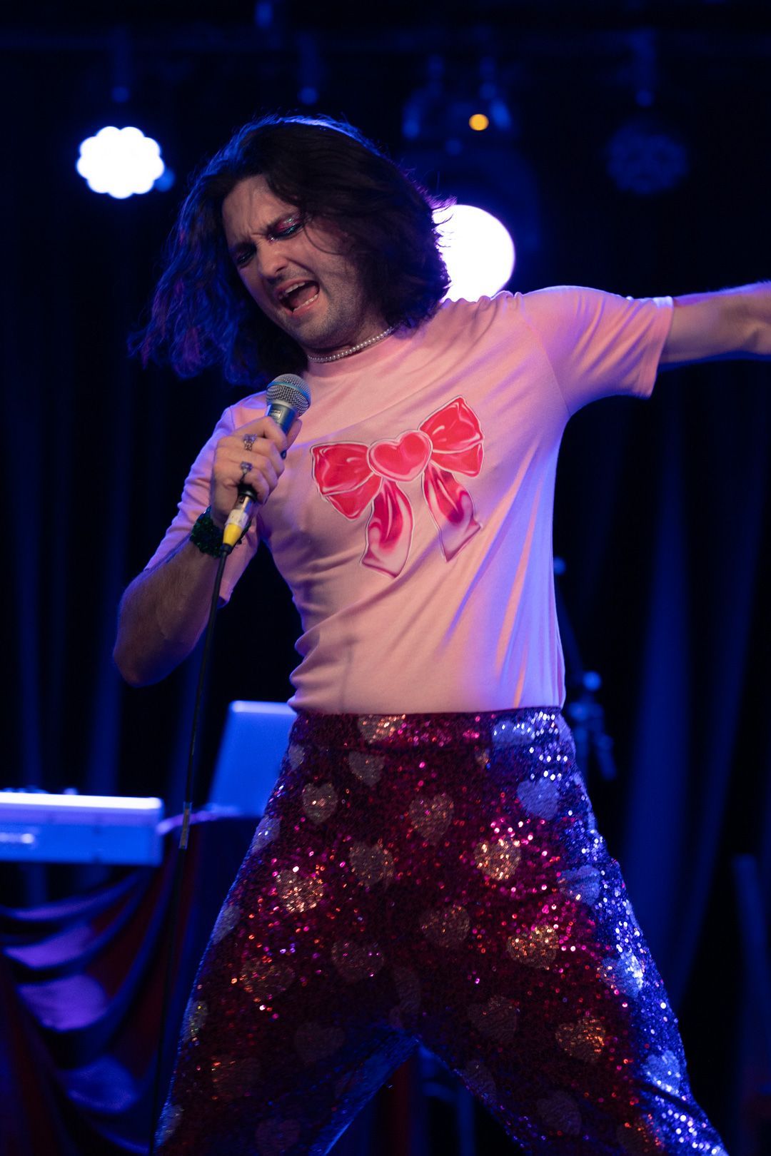 A man is singing into a microphone while wearing a pink shirt with a bow on it