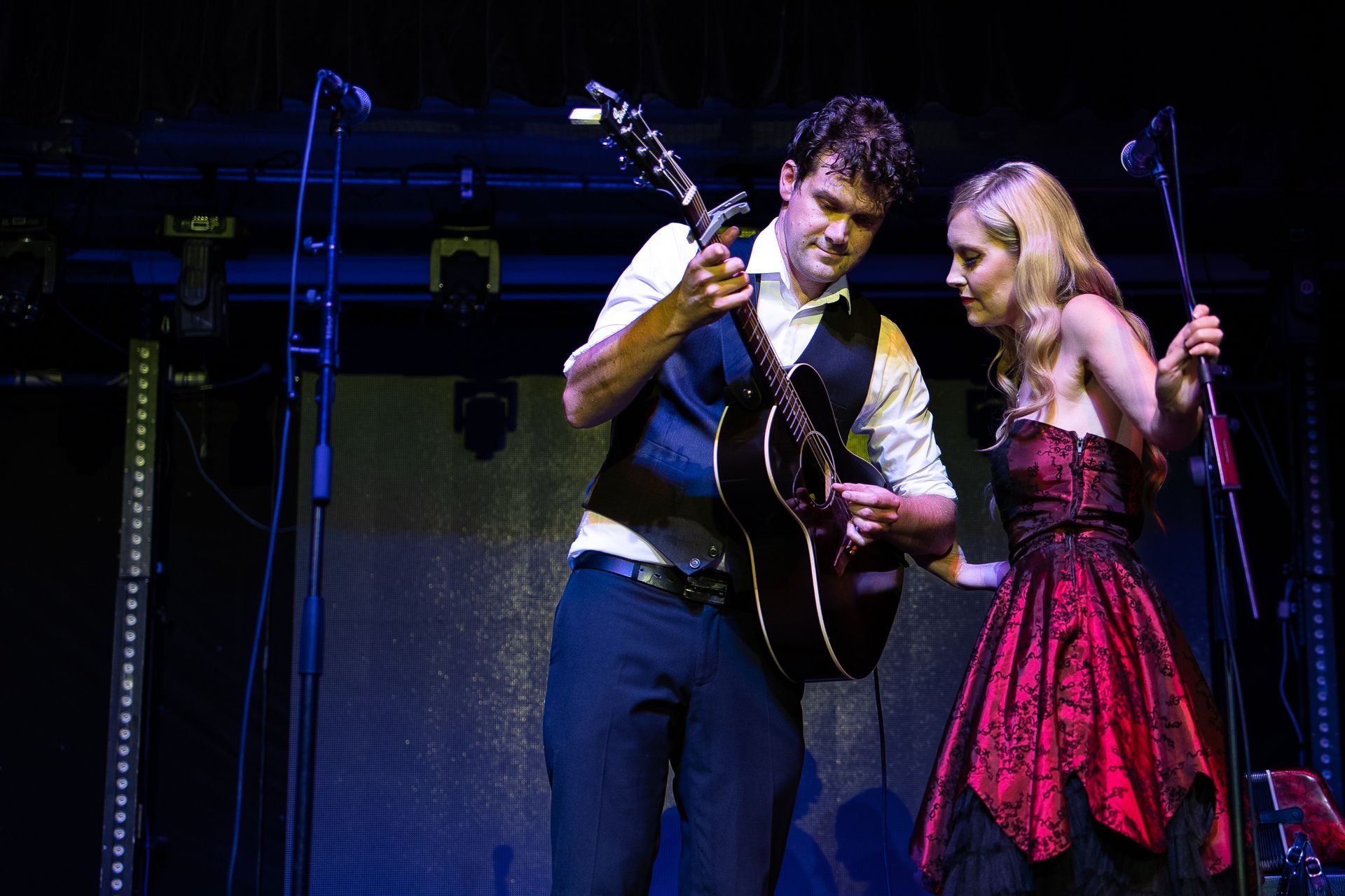 A man is playing a guitar next to a woman in a red dress on a stage.