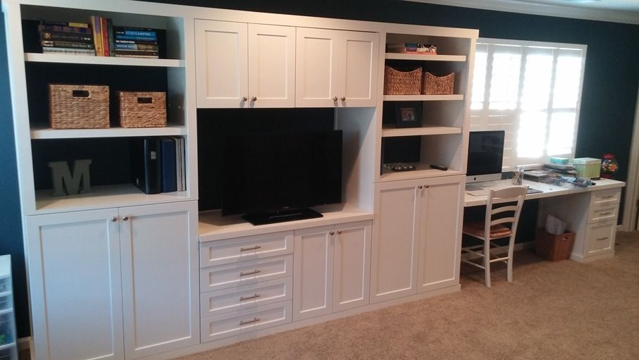 A room with white cabinets , shelves , a desk and a television.
