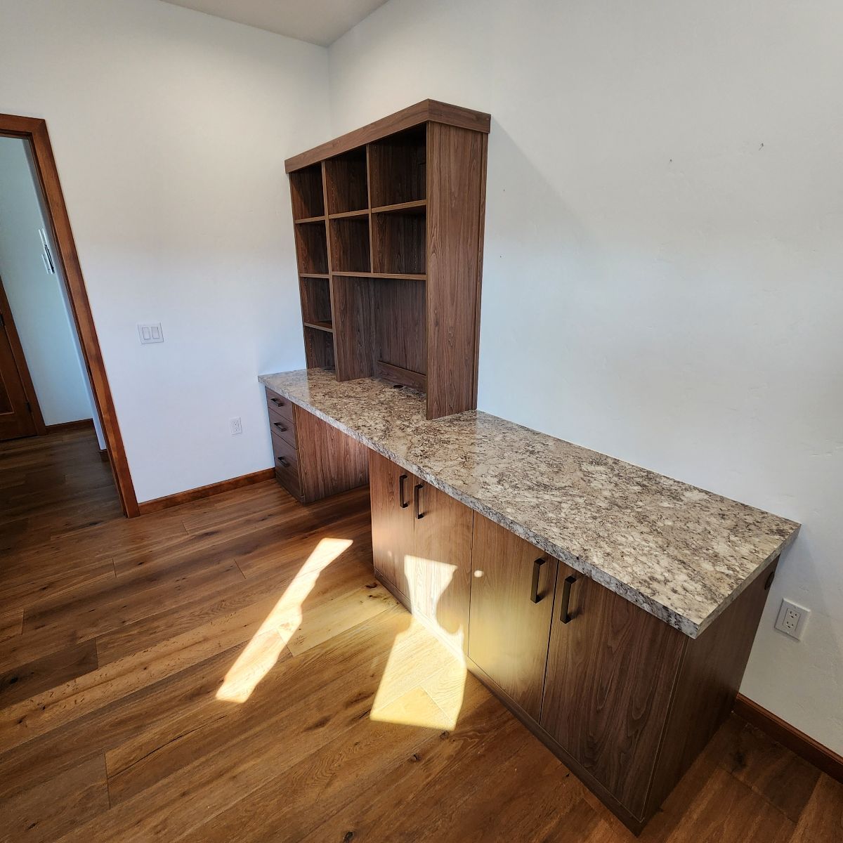 Built-in wooden desk with granite countertop and cubby shelves against a white wall in a room with wood flooring.