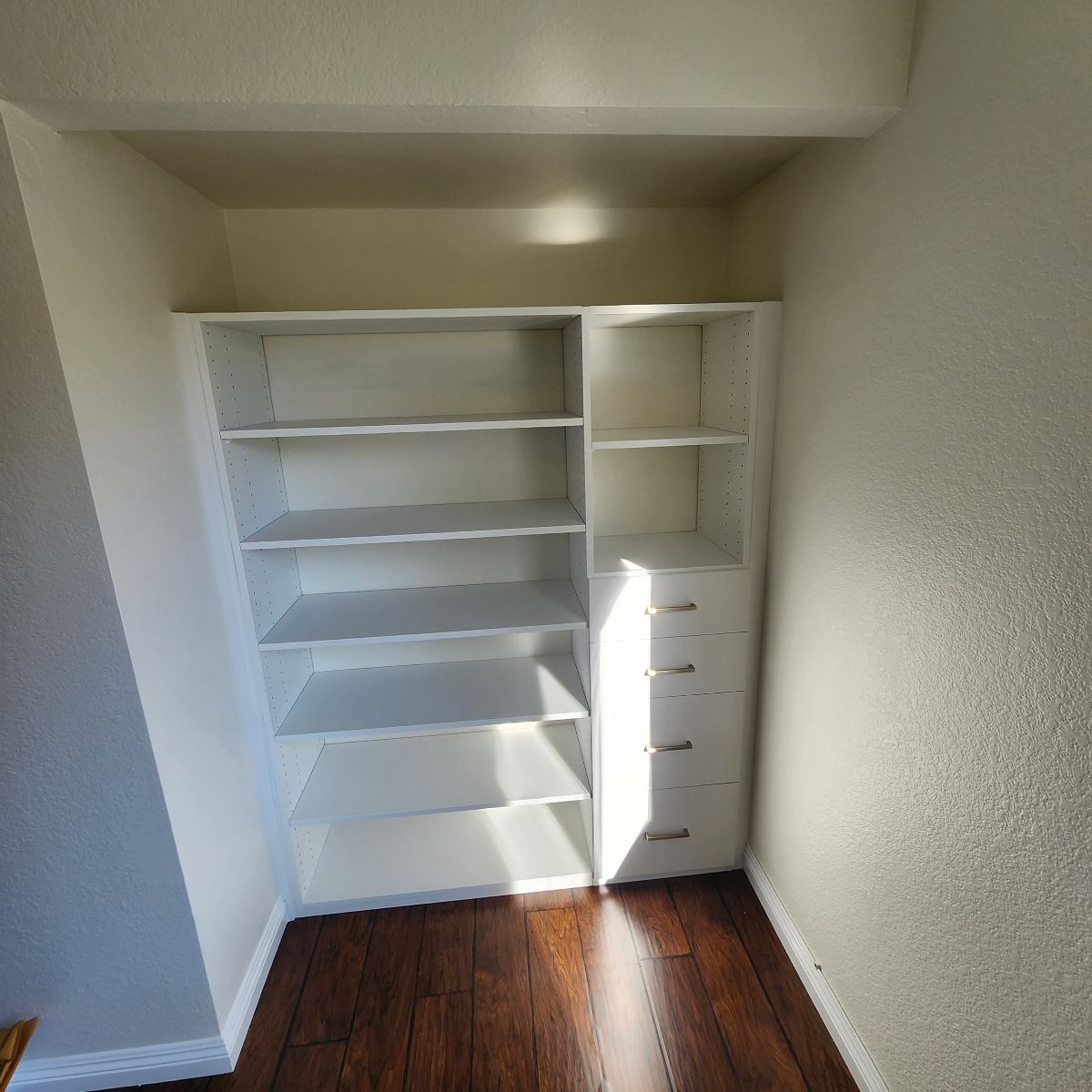 White built-in shelving unit with multiple shelves and drawers in a recessed space with a wood floor.