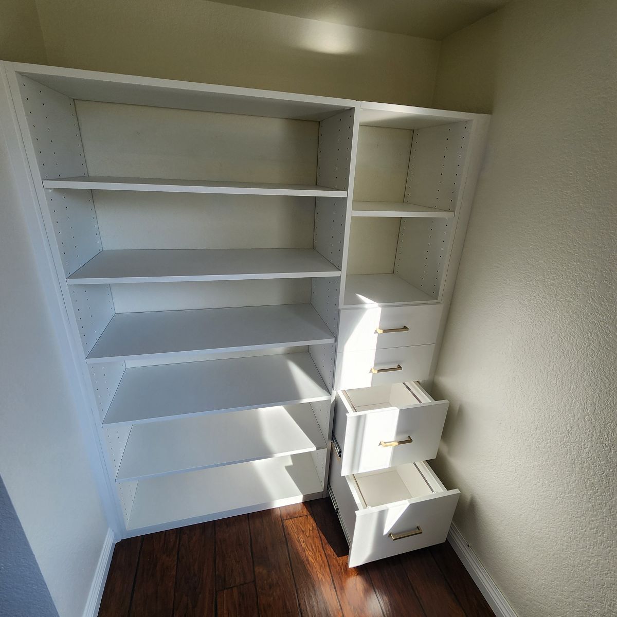 White built-in shelves in a corner of a room, with drawers and adjustable shelves, against a beige wall.
