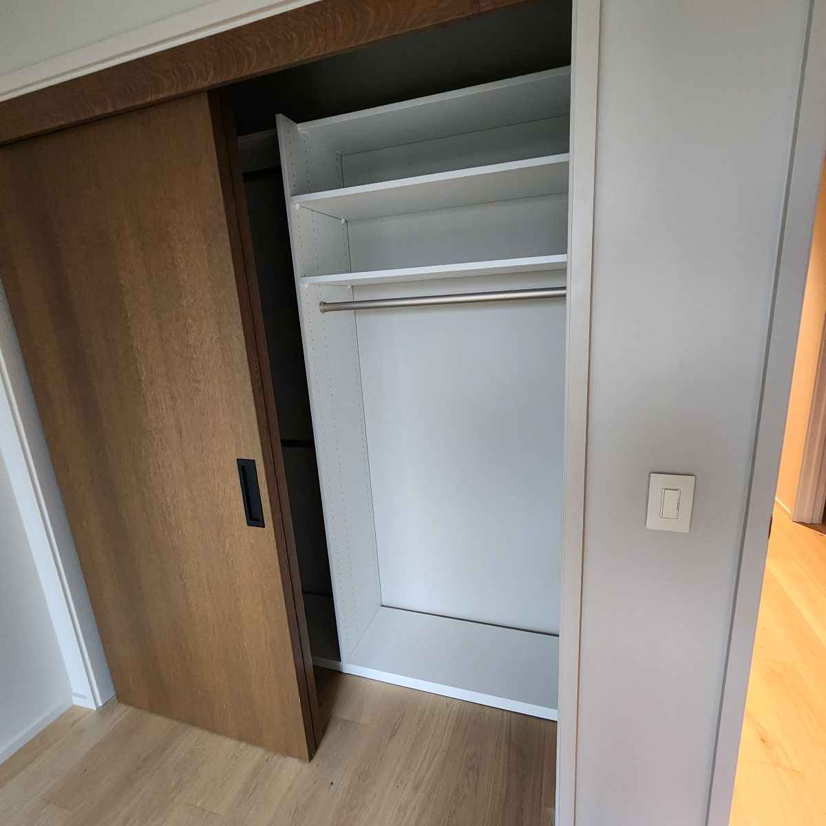 Open closet with sliding wooden door, shelves, and hanging rod. Light-colored wood and white interior.