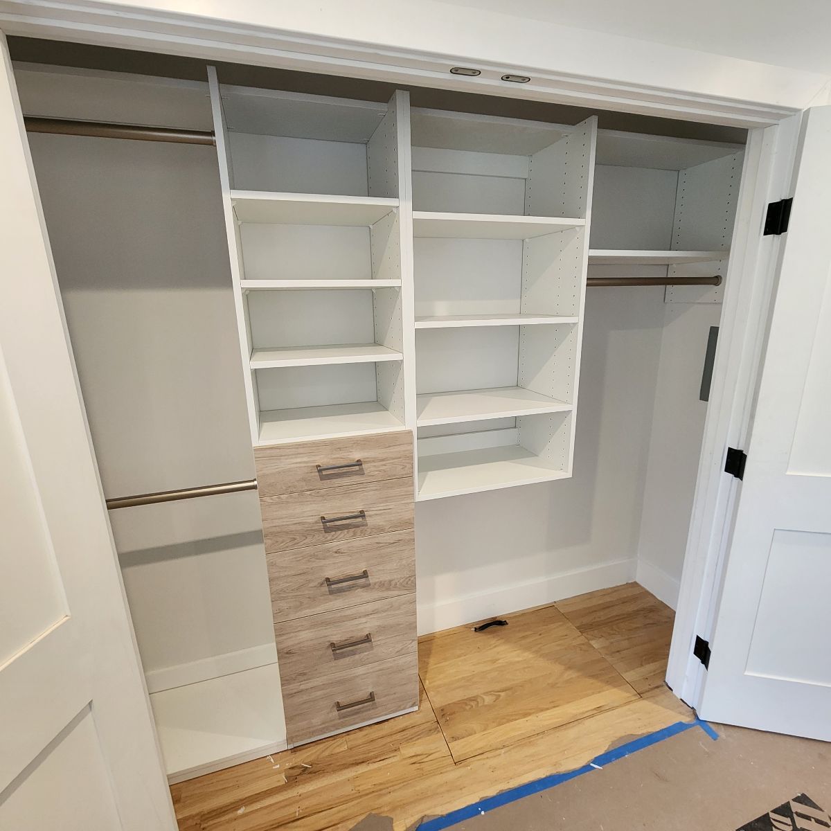 Closet interior with white shelves and drawers, and a hanging rod on both sides. The floor has light wood planks.