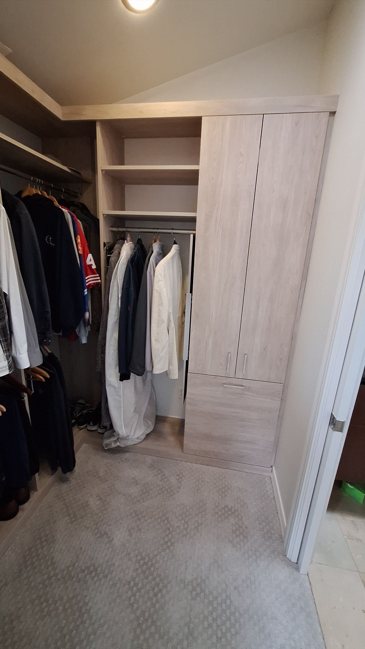 Walk-in closet with beige built-in shelves and rods, clothes hanging, light gray carpet and wooden door frame.
