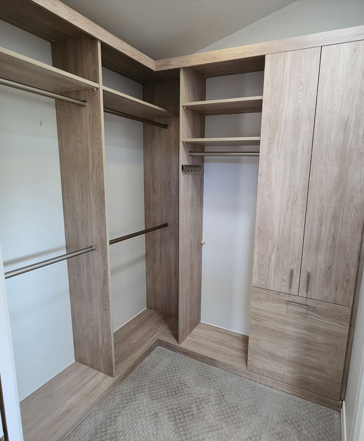 Corner walk-in closet with light wood shelving, rods, and closed cabinets against white walls, carpeted floor.