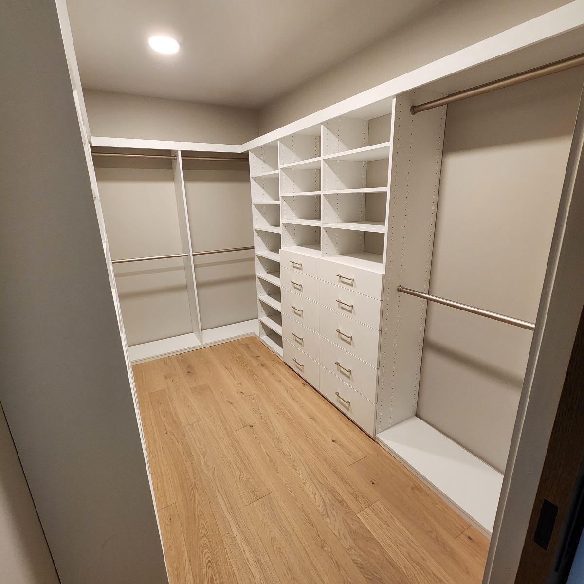 Walk-in closet with white shelving, drawers, and hanging rods. Light wood floor.
