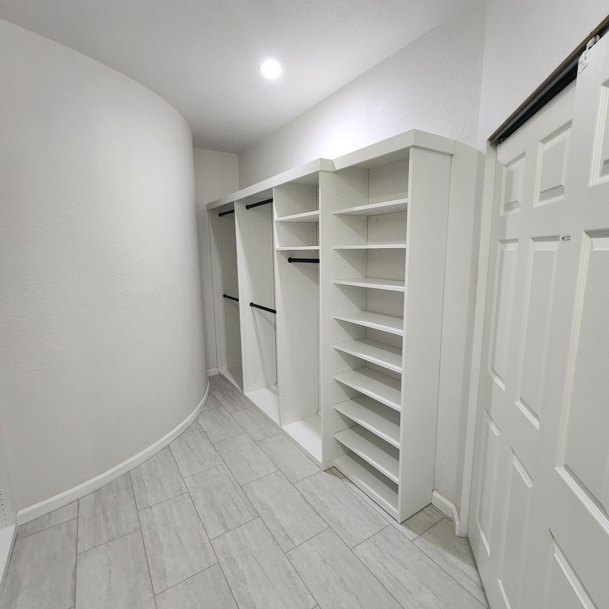 White walk-in closet with shelves, hanging rods, and a closed door. Gray tile floor.