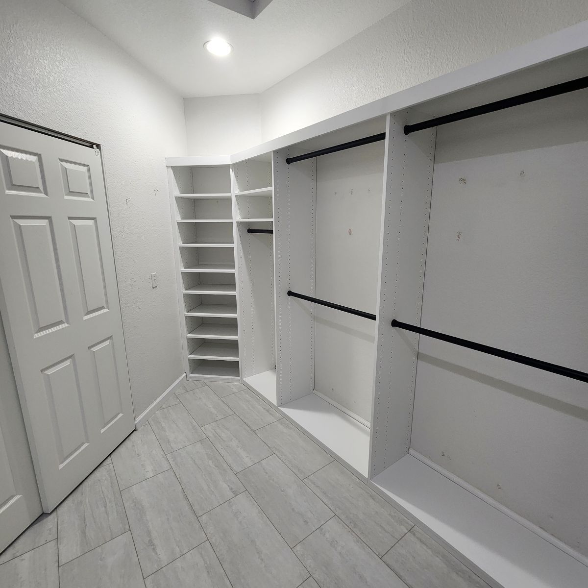 Empty white walk-in closet with shelves, hanging rods, and a closed white door; light gray tiled floor.