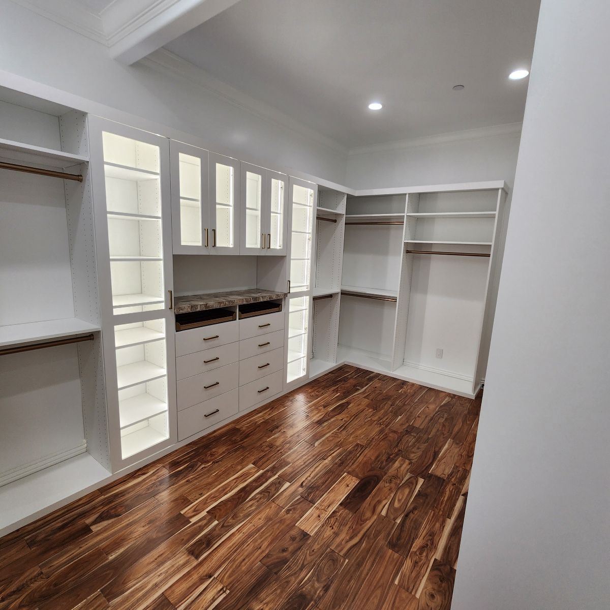 Spacious, white walk-in closet with wooden floors, built-in shelves, drawers, and glass-door cabinets.