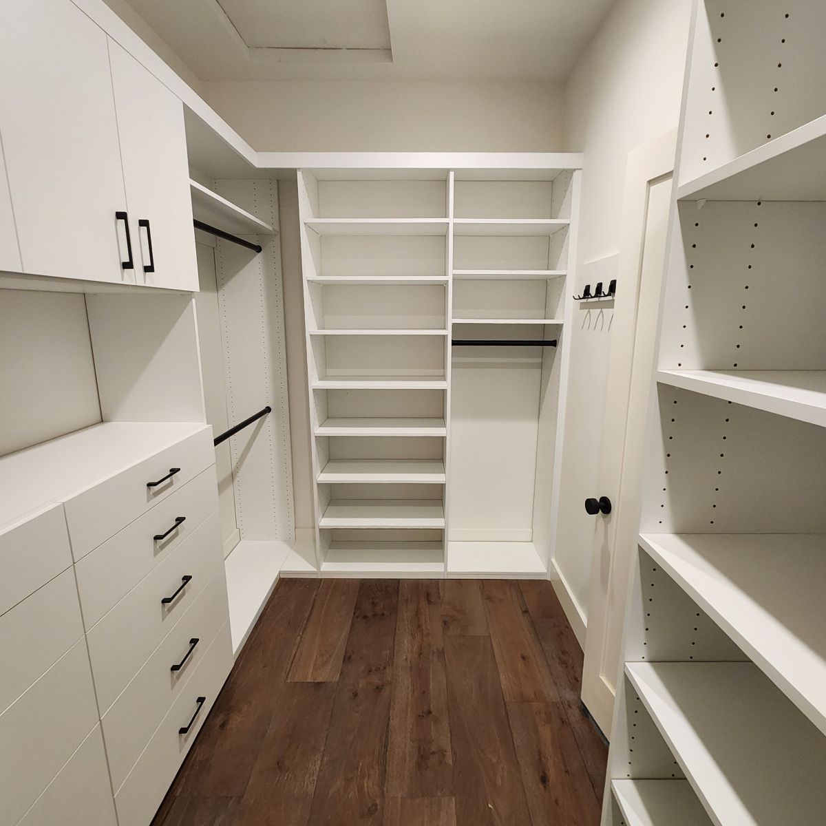 Walk-in closet with white shelves and cabinetry, wooden floor, open door.