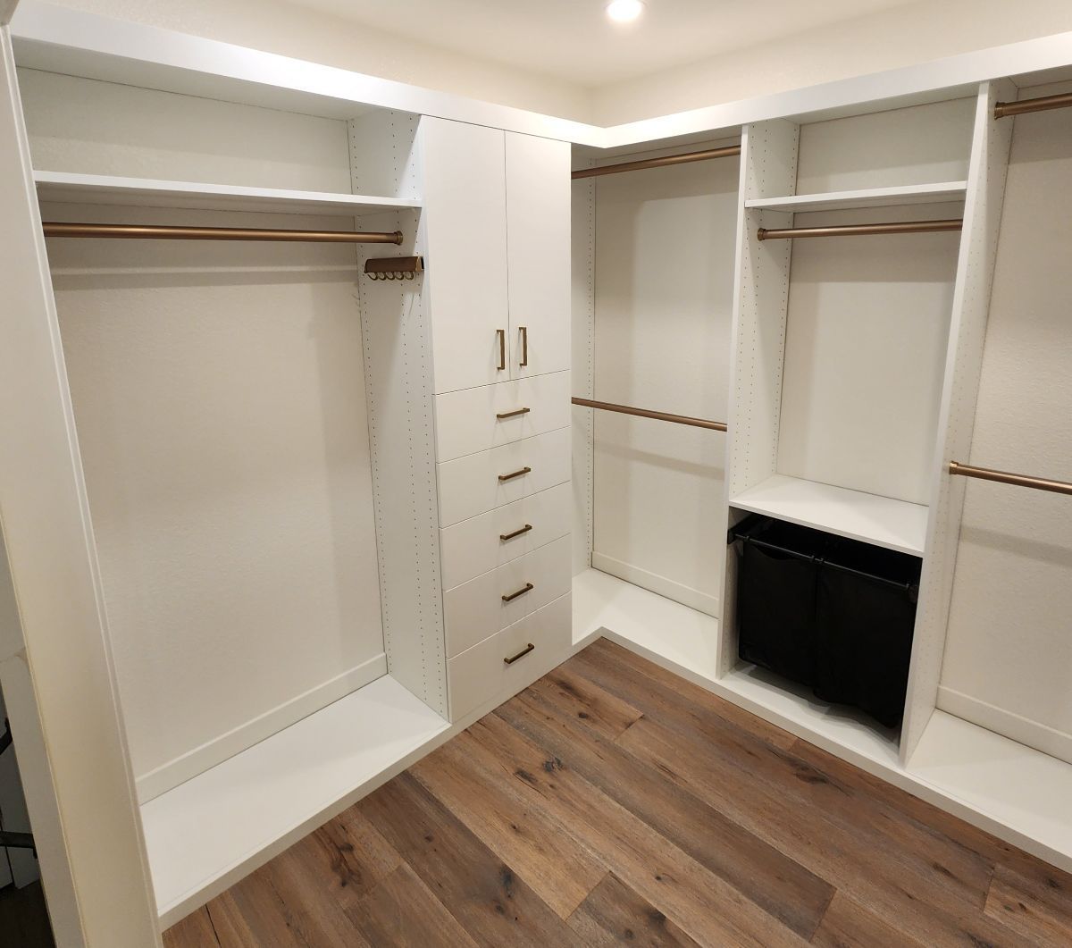 White walk-in closet with shelves, hanging rods, and a center tower with drawers. Wooden floor.