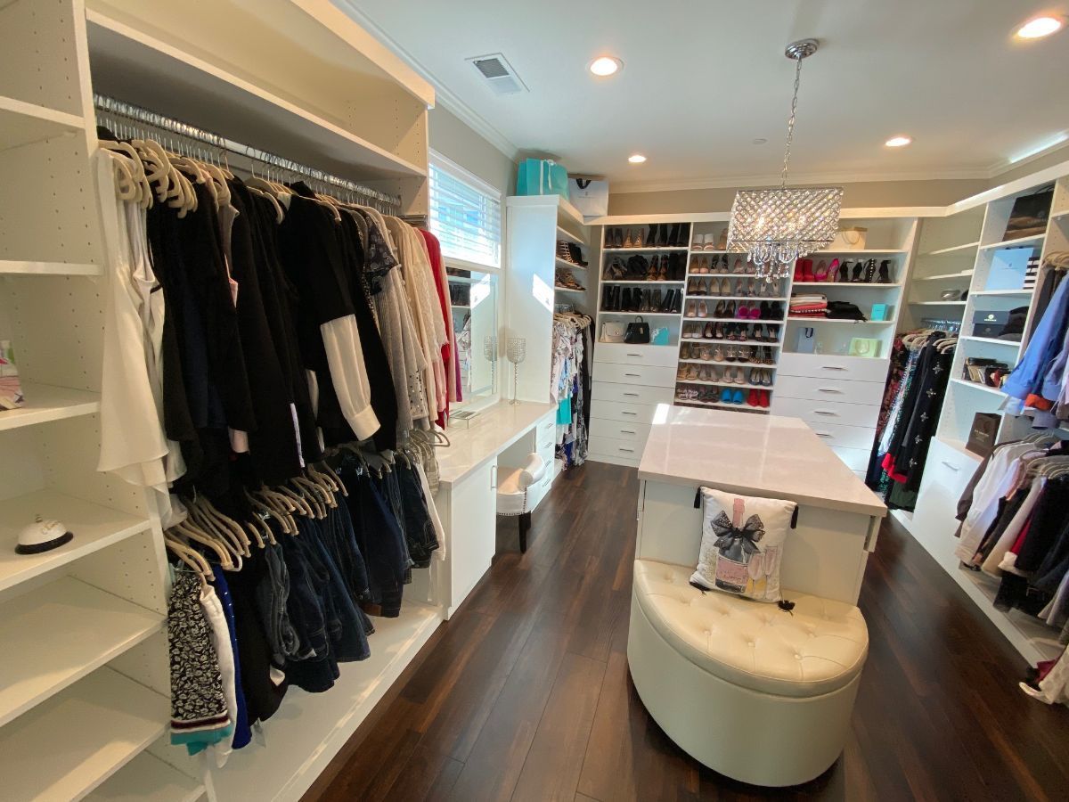 Walk-in closet with white shelves, hanging clothes, and dark wood floors. A center ottoman is present.