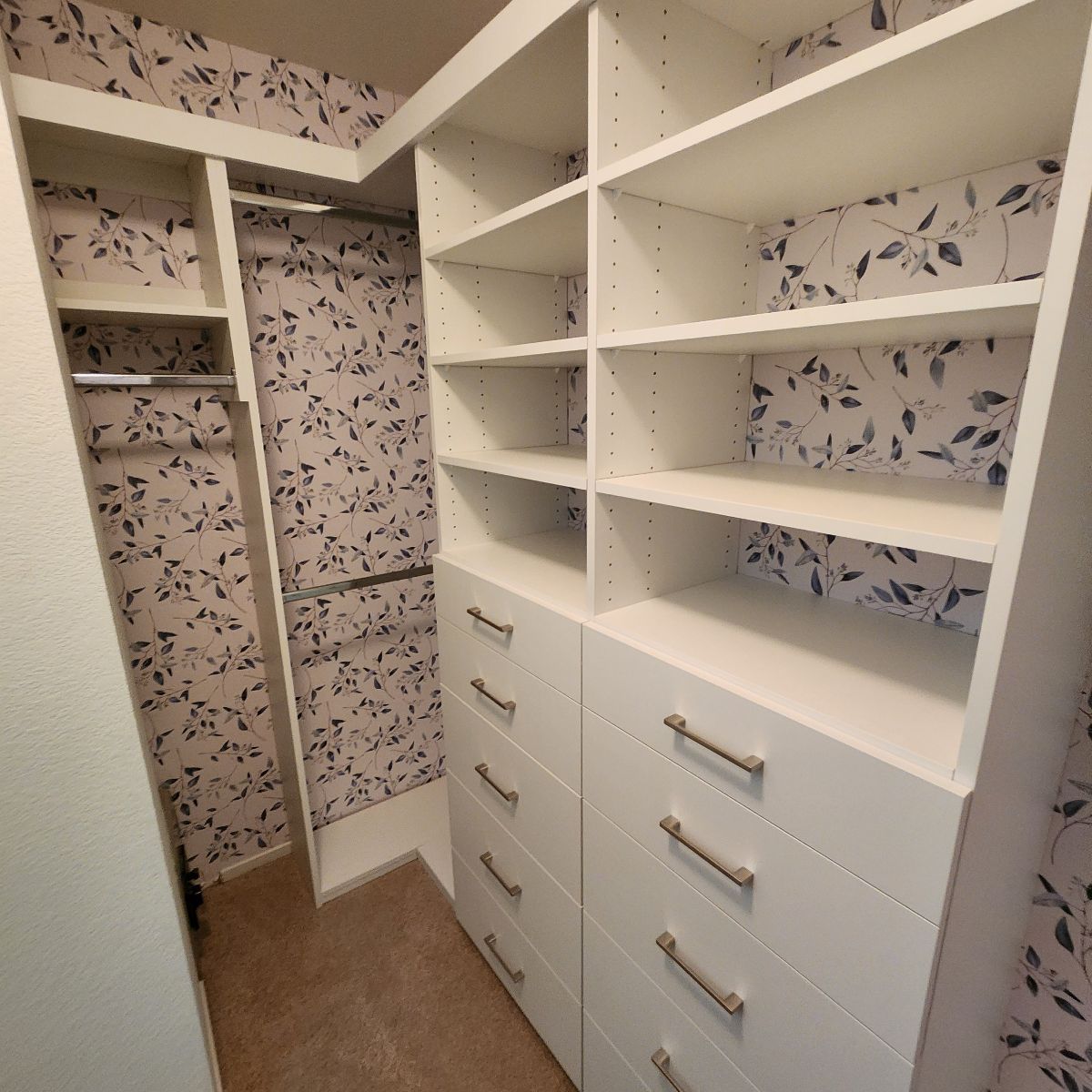 Walk-in closet with white shelves, drawers, and floral wallpaper.