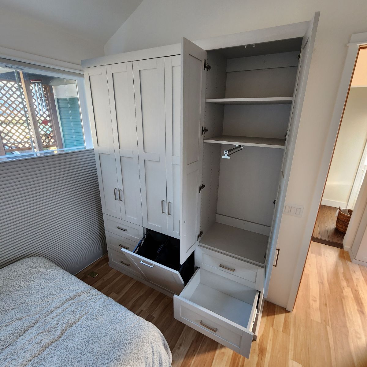 White wardrobe with open doors and drawers in a bedroom next to a bed and window.