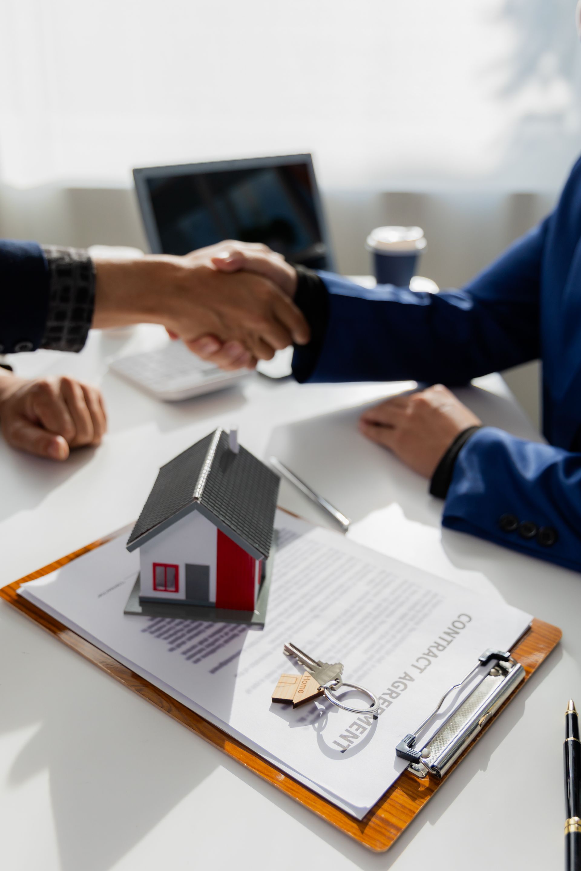 Two people are shaking hands over a clipboard with a model house and keys on it.