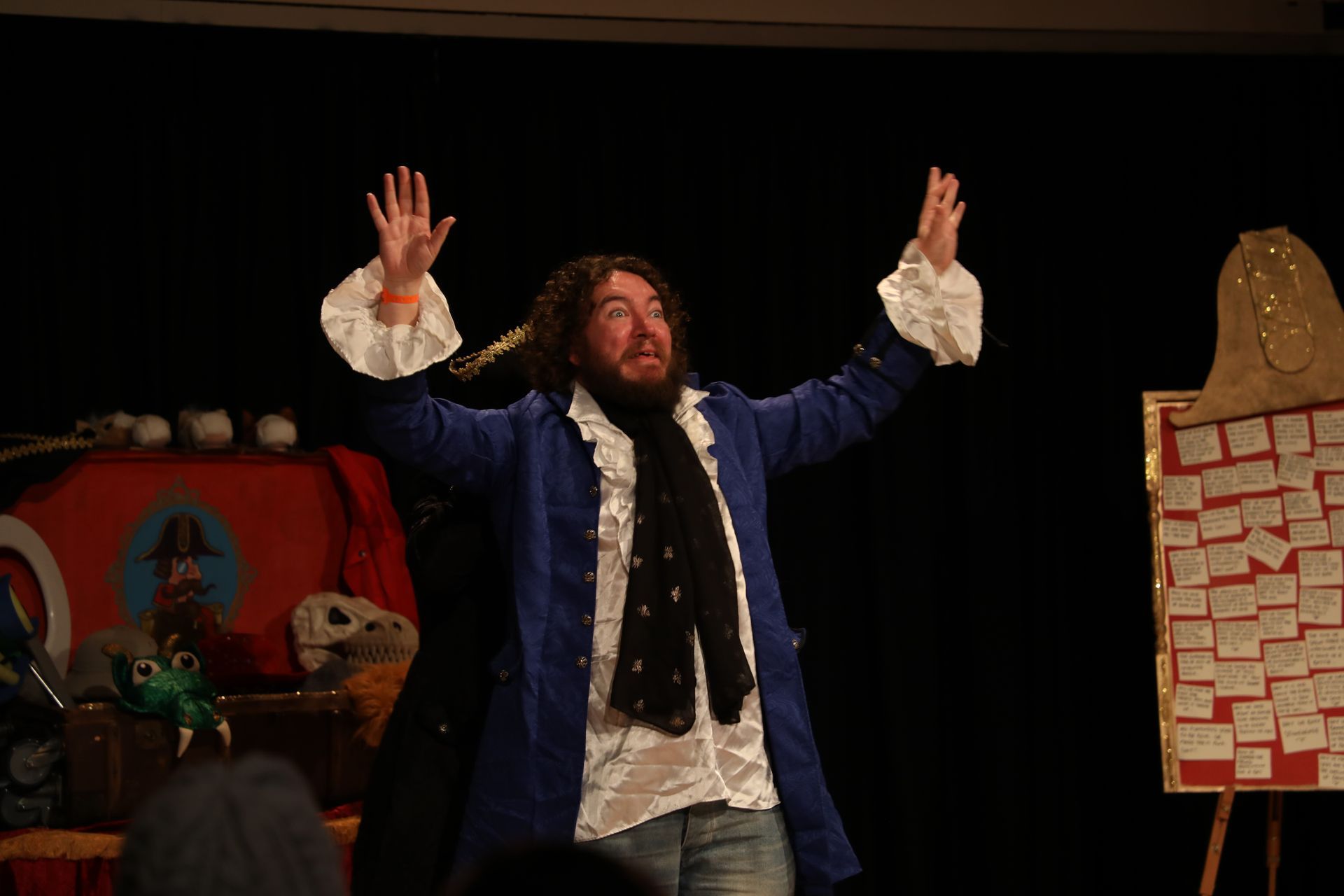 Man in blue coat with arms raised, onstage with props.
