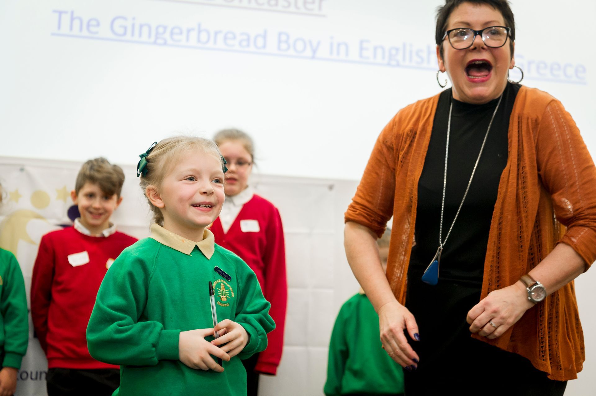 Woman with glasses and students in green sweaters on a stage; a screen shows