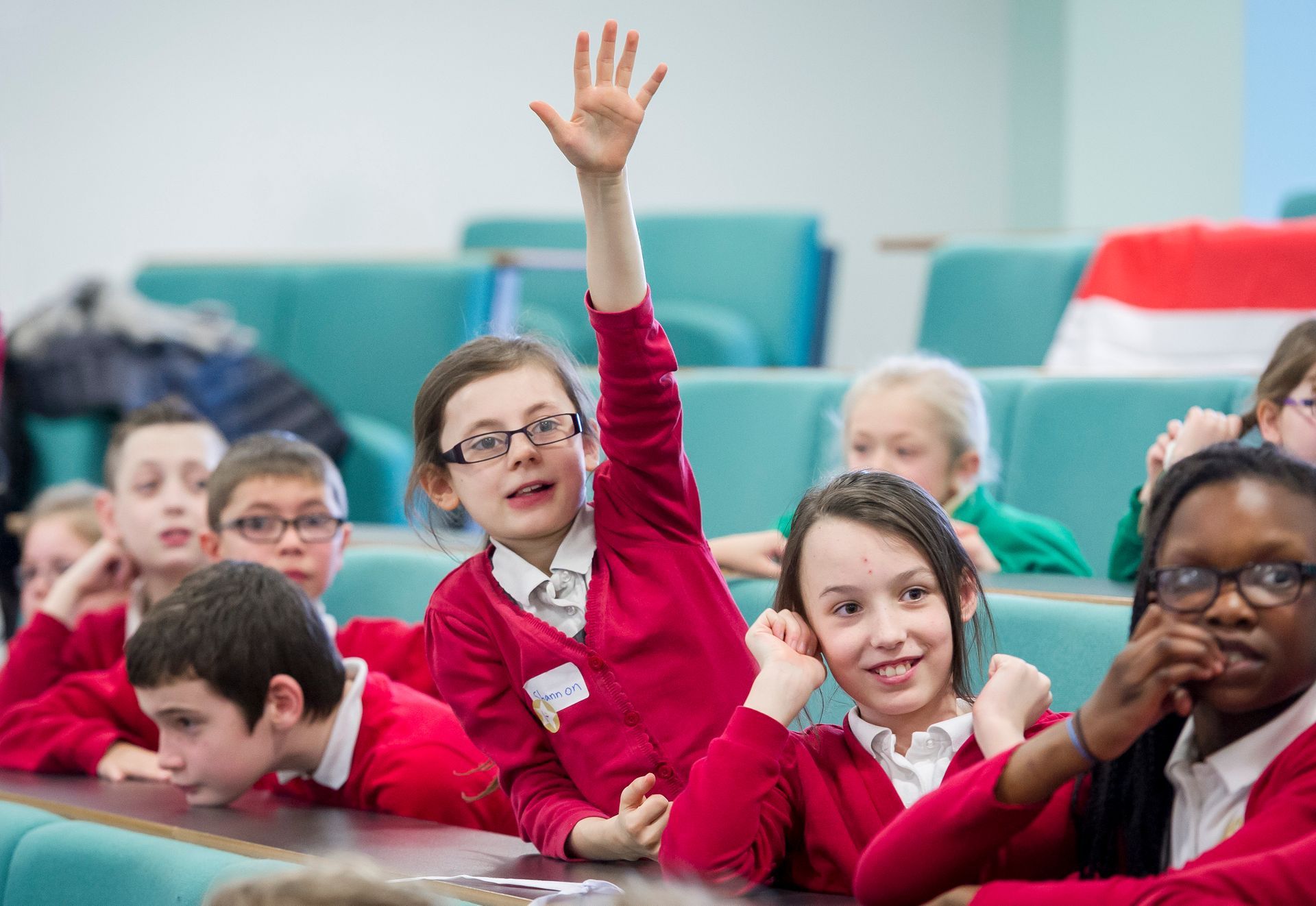 Group of schoolchildren in red uniforms, one raising their hand in a lecture hall.