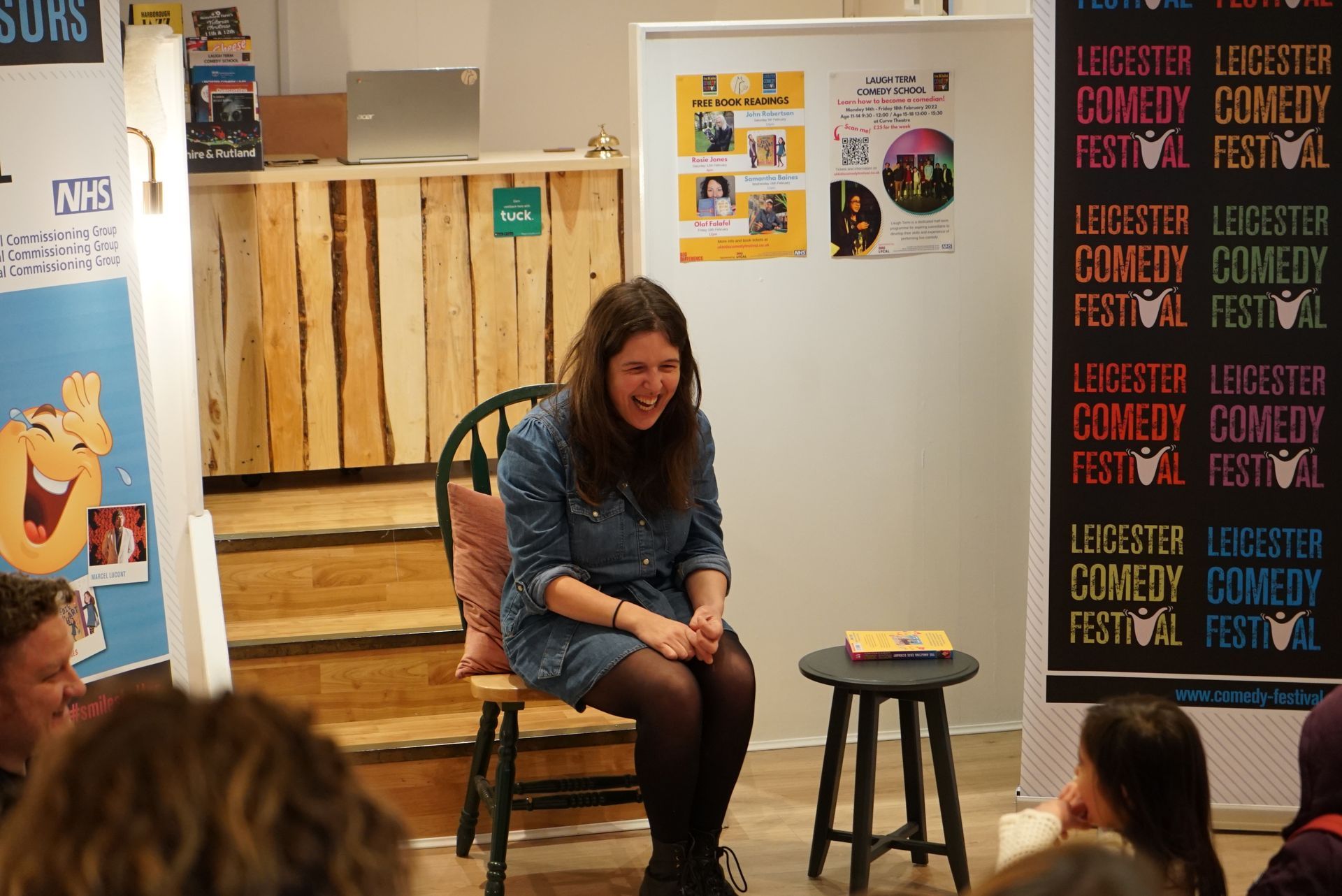 Woman laughing, seated on a stage, at a comedy festival with posters in the background.