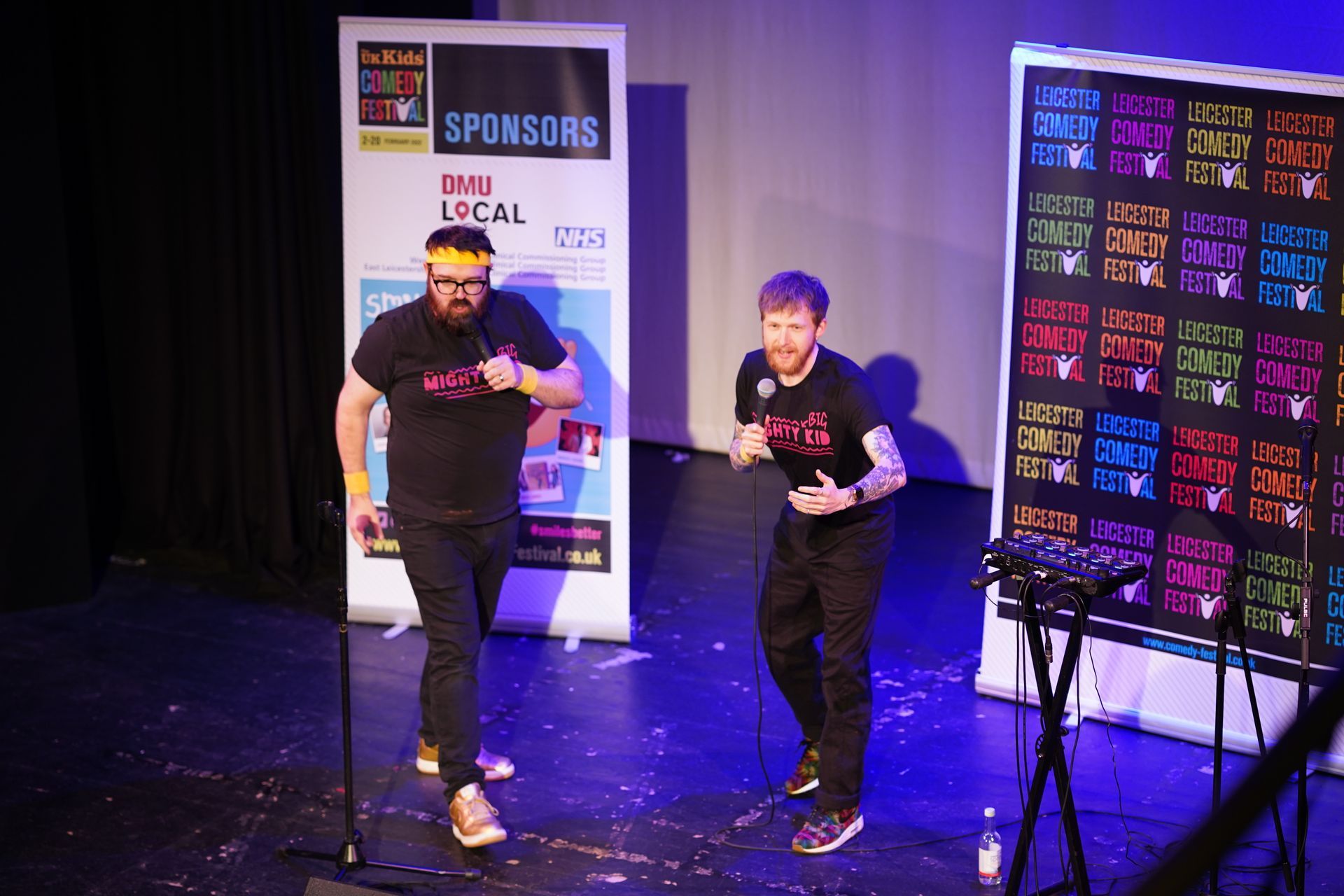 Two people on a stage with a presentation screen and banner. The setting is a theater.
