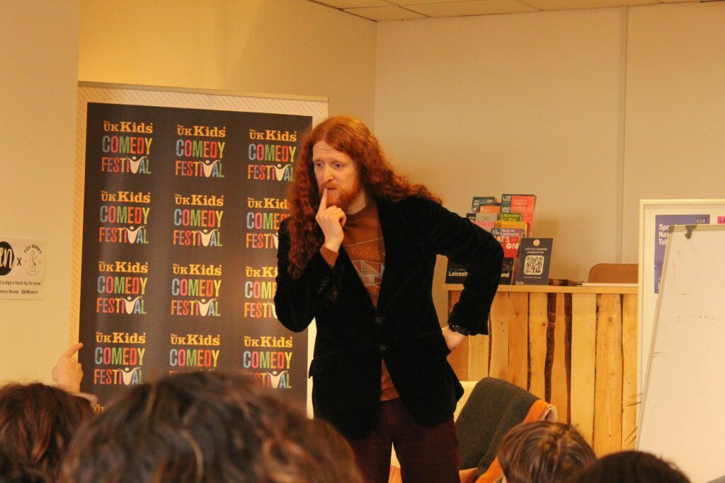 A person with long red hair wearing a dark jacket and burgundy pants on stage at a comedy festival.