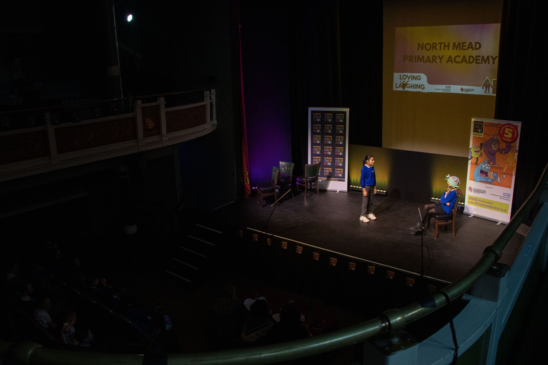 Two people on a stage with a presentation screen and banner. The setting is a theater.