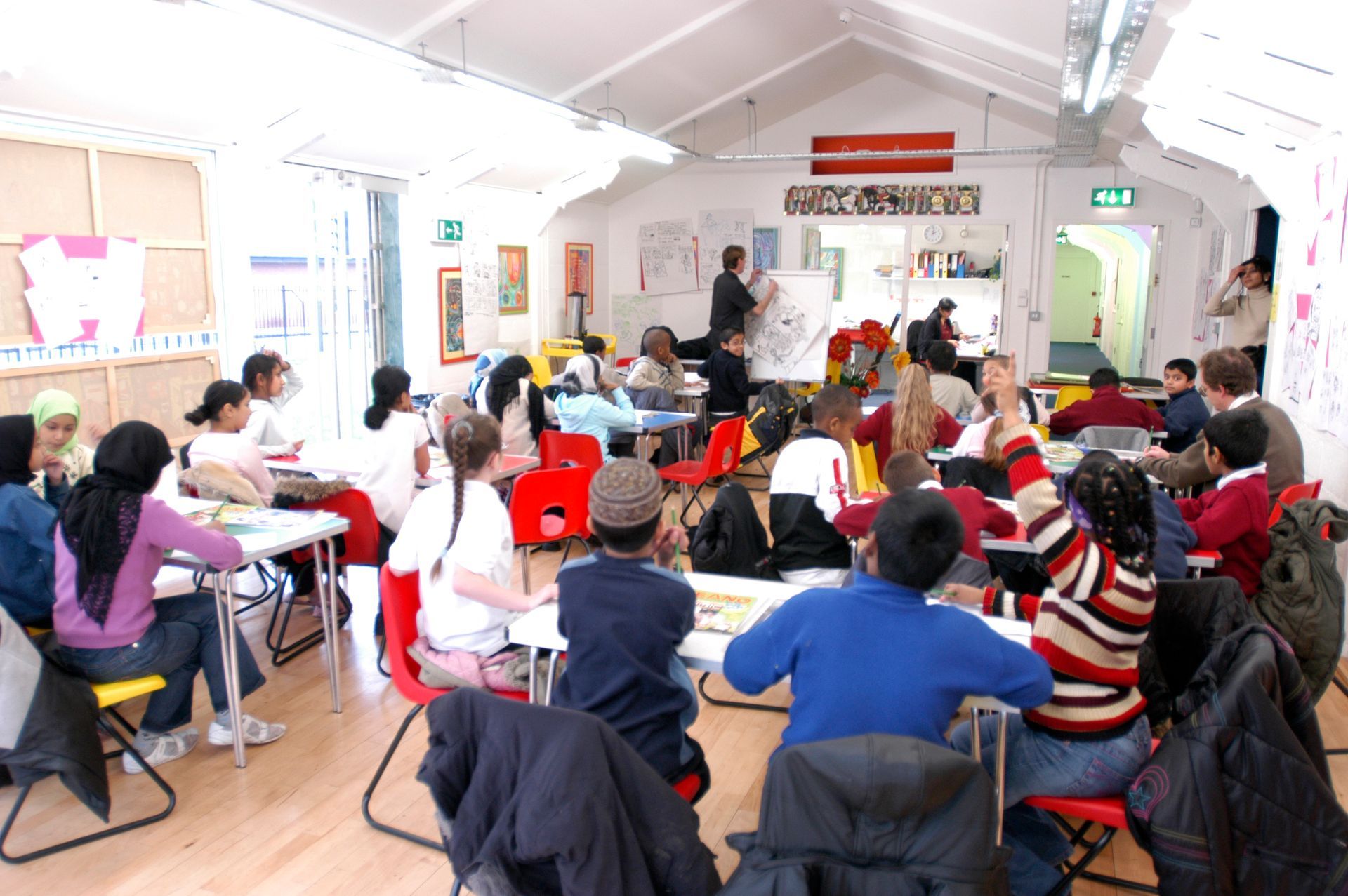 Children in a brightly lit classroom, some raising hands, with a teacher at the front.