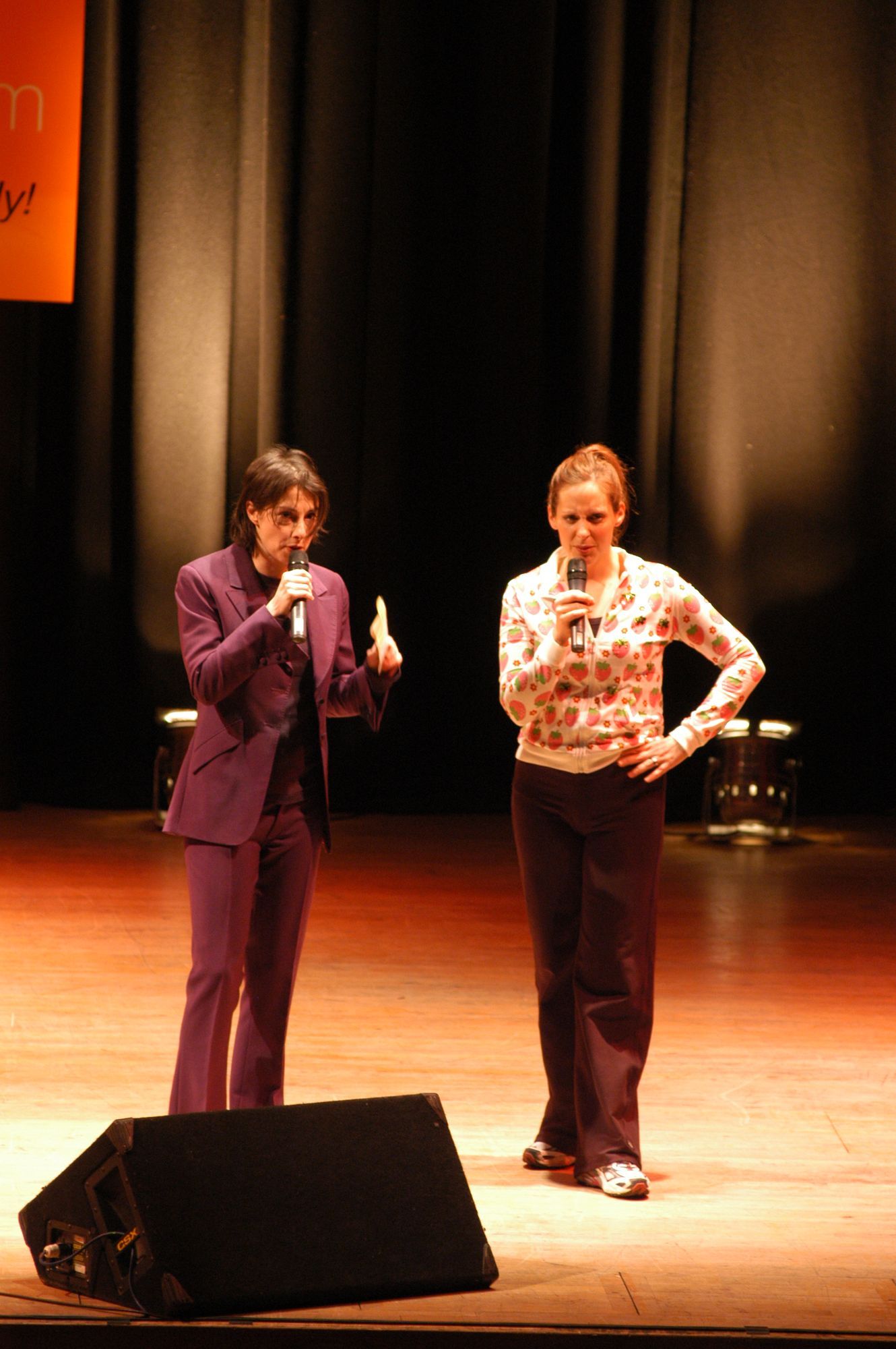 Two people on a stage with microphones: one in a purple suit gestures, the other in a patterned shirt stands beside.