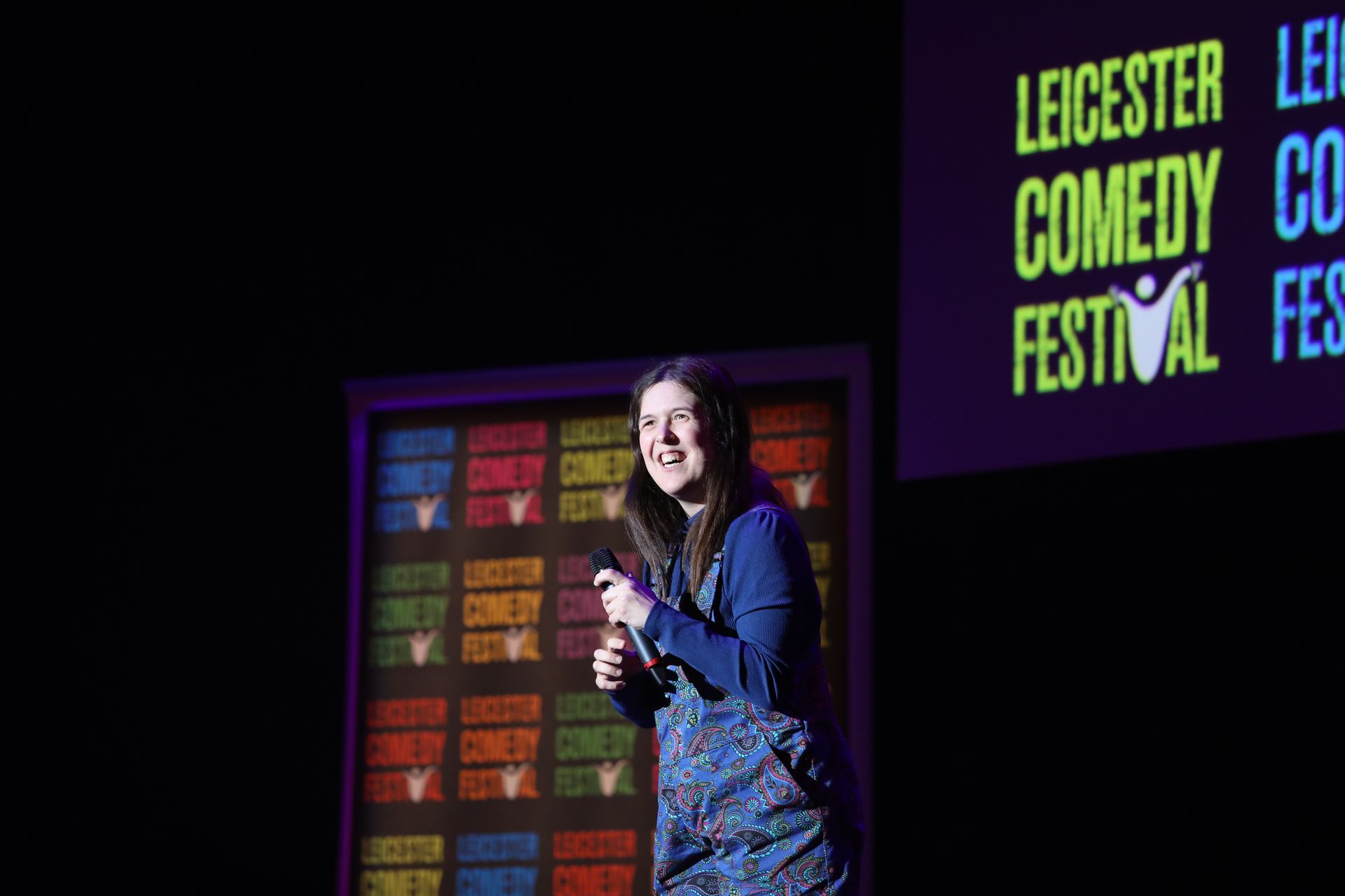 Woman on stage at Leicester Comedy Festival, holding a microphone, smiling.