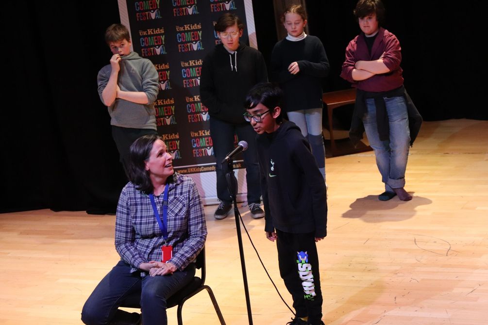 Woman with blonde hair, wearing a floral top, seated at a table.