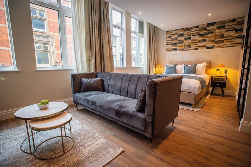 Hotel suite with velvet sofa, bed, side tables, large windows, and decorative wall.