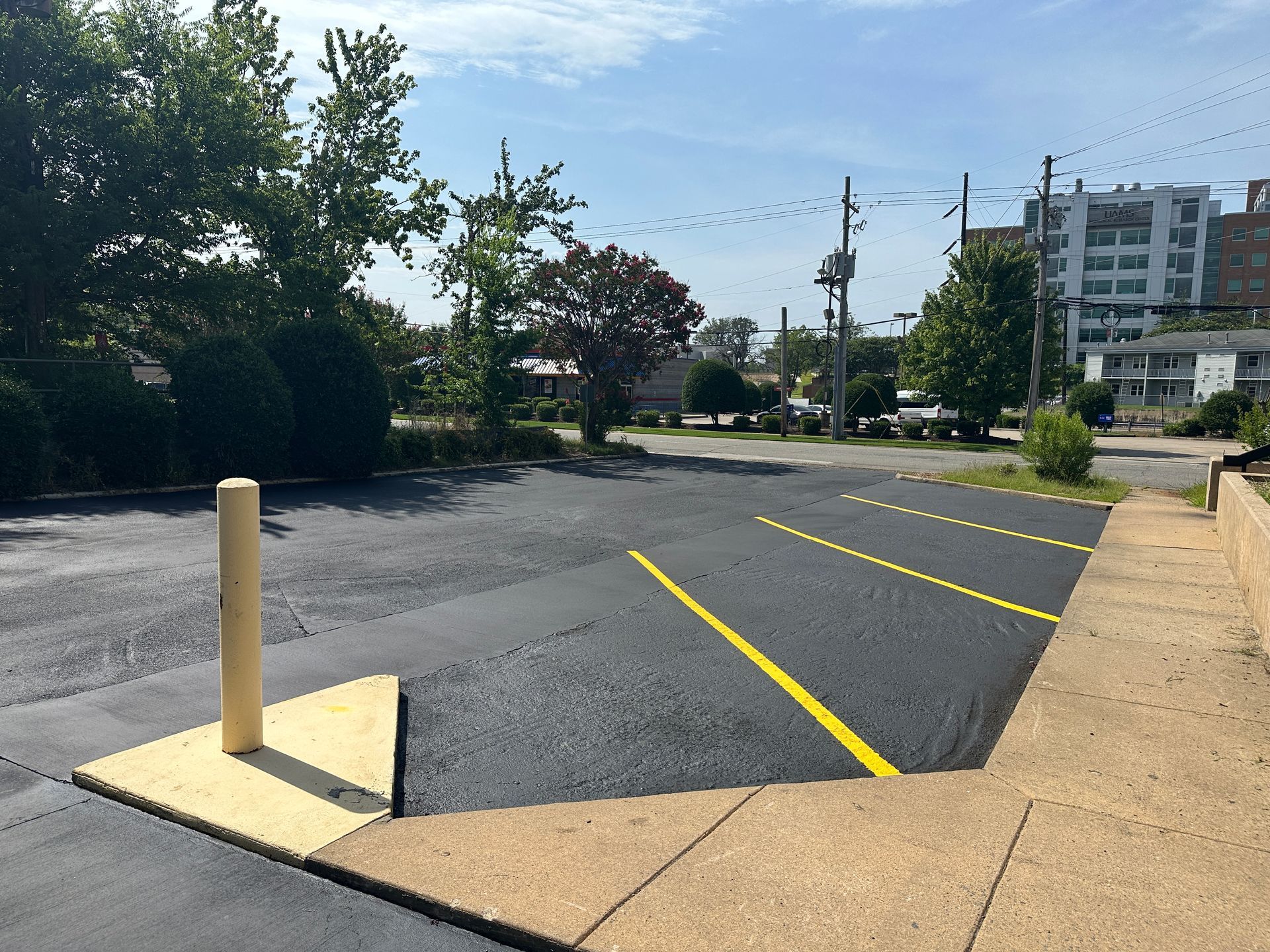 Newly paved parking lot with yellow lines, a concrete border, and a tan bollard on a sunny day.