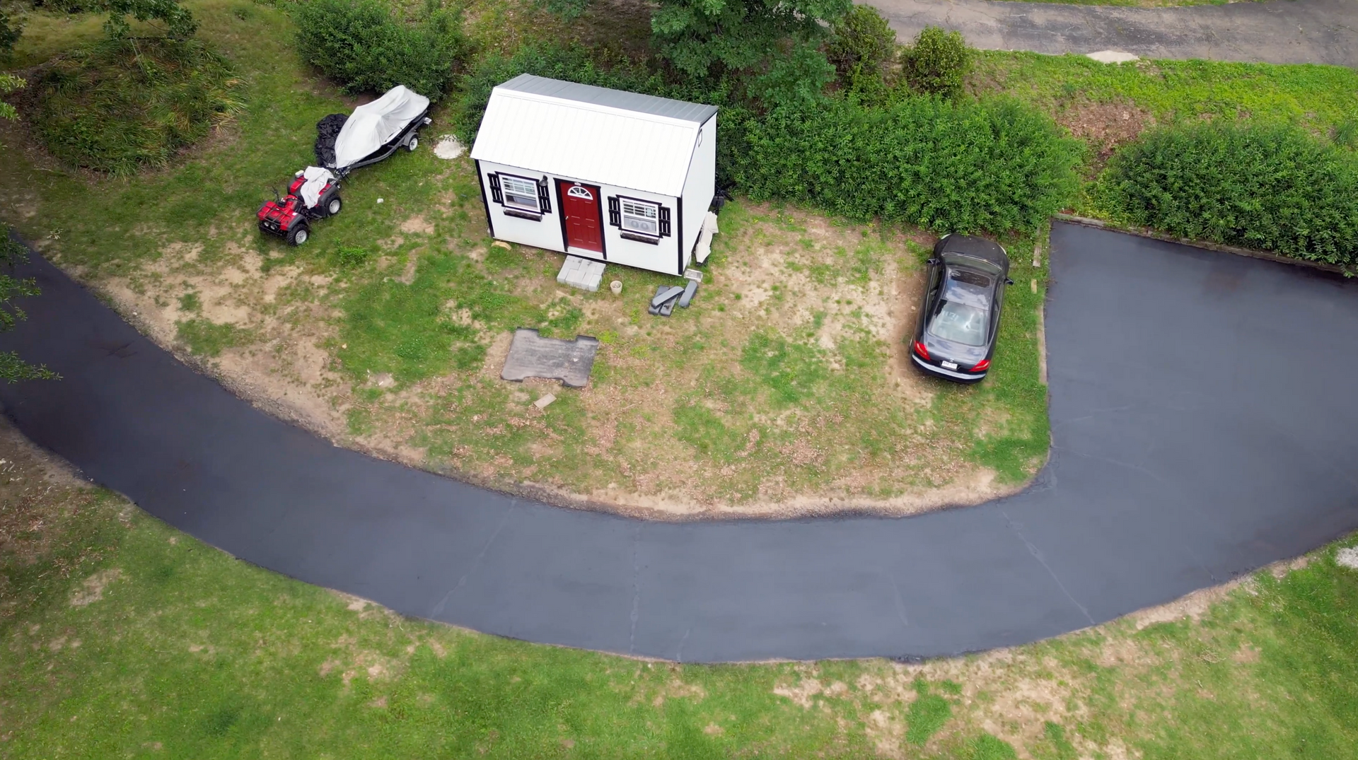 Aerial view of small white building, black paved driveway, car, boat, and shrubs.