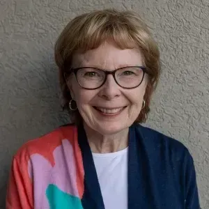 Woman with glasses, smiling, wearing a colorful sweater and a white shirt against a textured wall.