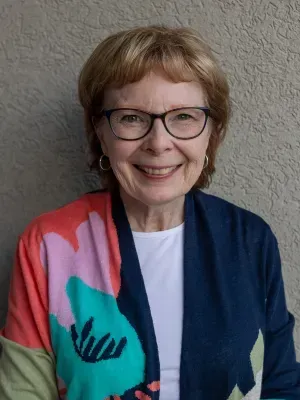 Woman with glasses, smiling, wearing colorful floral cardigan and white top against a neutral wall.