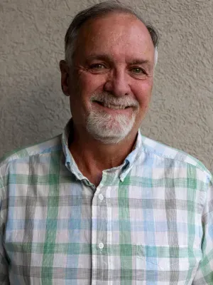 Smiling older man with a gray beard, wearing a plaid shirt against a neutral background.